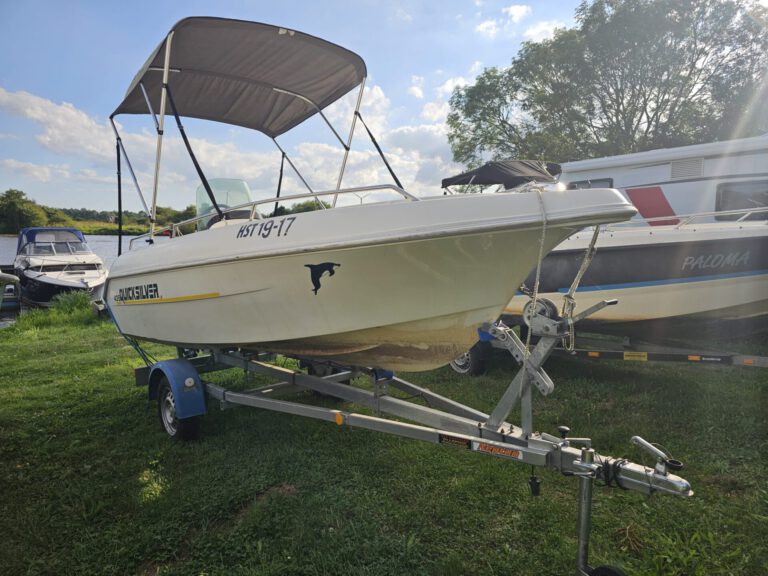Weser-Vermietung: Wohnmobil, Boot & Motorrad mieten an der Weser 1 Weißes Quicksilver Motorboot mit grauem Sonnendach und Trailer, bereit zur Vermietung, geparkt auf grünem Gras neben weiteren Booten und einem Wohnmobil.