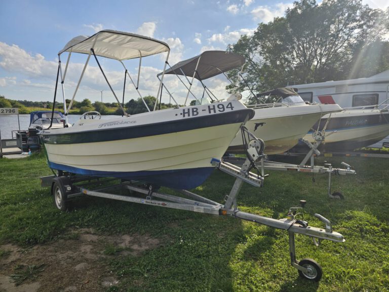 Weser-Vermietung: Wohnmobil, Boot & Motorrad mieten an der Weser 6 Beiges Motorboot "Francesca" mit blauem Rumpf und Sonnendach auf einem Anhänger, daneben weitere Boote und ein Wohnmobil, bereit zur Vermietung.