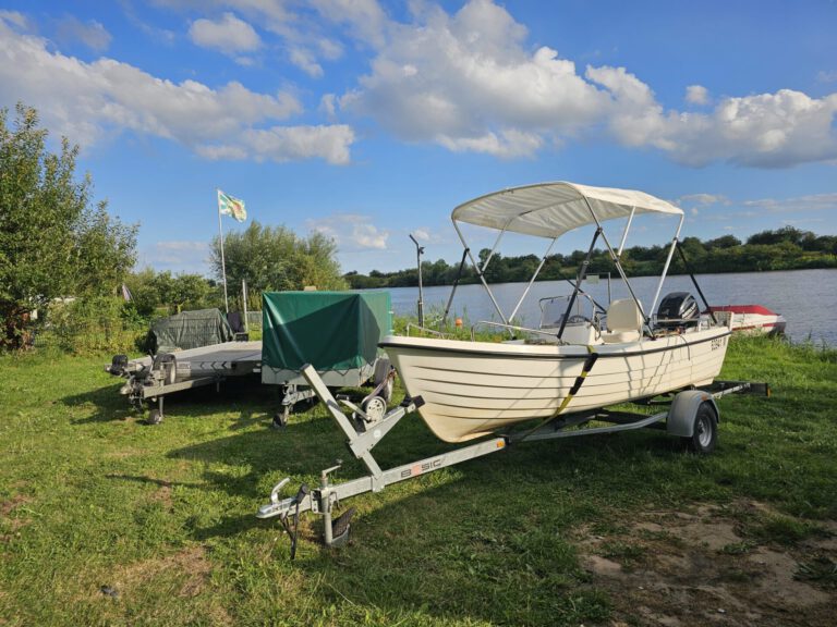 Weser-Vermietung: Wohnmobil, Boot & Motorrad mieten an der Weser 4 Helles Ruder- oder Motorboot mit Sonnendach auf einem Trailer, daneben ein zweiter Bootsanhänger mit grüner Plane, alles auf einer Wiese am Wasser.