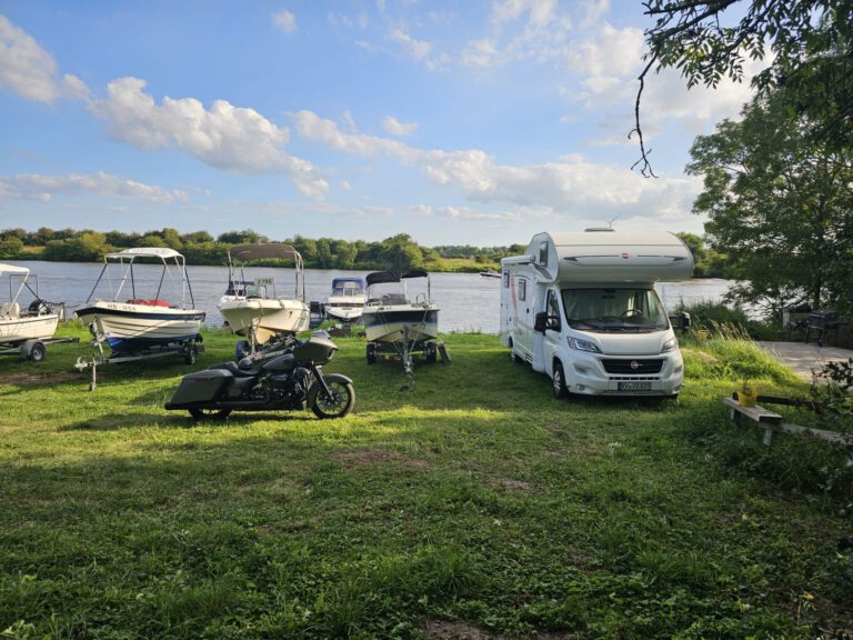 Weser-Vermietung: Wohnmobil, Boot & Motorrad mieten an der Weser 12 Weißer Fiat Ducato 6-Personen-Wohnmobil, schwarzes Motorrad und mehrere Boote auf Anhängern am Ufer der Weser an einem sonnigen Tag. Mieten Sie Ihr Abenteuer bei weser-vermietung.