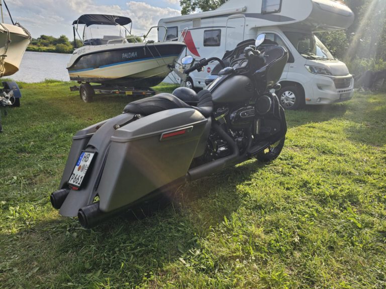 Weser-Vermietung: Wohnmobil, Boot & Motorrad mieten an der Weser 10 Mattschwarze Harley-Davidson Street Glide mit großem Koffer am Heck, geparkt auf grünem Gras neben einem weißen Wohnmobil und einem Boot mit dem Namen "PALOMA" auf einem Anhänger am Ufer der Weser.
