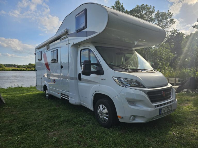 Weser-Vermietung: Wohnmobil, Boot & Motorrad mieten an der Weser 2 Modernes, weißes Alkoven-Wohnmobil (vermutlich Fiat Ducato Basis) mit roten und grauen Akzenten, geparkt auf einer grünen Wiese direkt am Ufer eines Flusses.